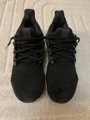Svarta Adidas Ultra Boost sneakers - Säljer ett par svarta Adidas Ultra Boost sneakers med stickad ovandel och klassiska tre ränder i plast på sidorna. Skorna har svart snörning och en vit, dämpad sula med svart yttersula. Perfekta för dig som gillar sportig och stilren look. Priset kan diskuteras vid snabb affär.