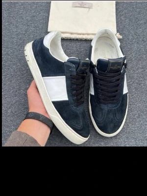 Valentino Garavani svarta sneakers - Snygga sneakers från Valentino Garavani i svart mocka med vita detaljer och vit sula. Klassisk låg modell med svarta skosnören och diskret logga på plösen. Perfekta för dig som vill ha en stilren och exklusiv look.storleken är 41 men passar 41-42