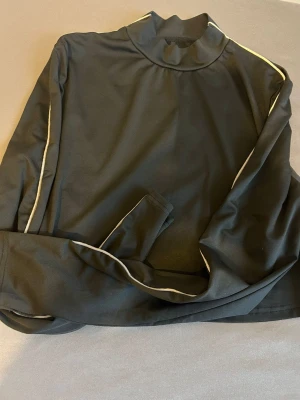 Svart träningströja med vita detaljer - Snygg svart långärmad träningströja med vita kontrastlinjer längs ärmarna. Tillverkad i ett stretchigt funktionsmaterial som andas, perfekt för löpning eller gym. Hög krage för extra komfort och stilren look.