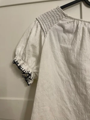 Vit blus med svarta detaljer - Säljer en luftig vit blus med korta puffärmar och svarta broderade detaljer vid ärmslut, axlar och nederkant. Blusen har en avslappnad passform och är tillverkad i ett krinklat bomullstyg. Perfekt för en trendig och avslappnad look.