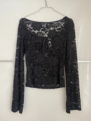 Svart spetsblus med snörning - Trendig svart långärmad blus från Pieces i genombruten spets med blommönster. Toppen har en rundad urringning och dekorativ snörning framtill. 