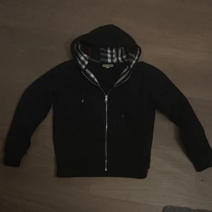 Svart Burberry hoodie med dragkedja - Snygg svart hoodie från Burberry med klassiskt rutigt foder i huvan och dragkedja framtill. Ikoniska Burberry-loggan är diskret broderad på bröstet. Perfekt för dig som gillar stilrena och exklusiva plagg. Tillverkad i mjukt bomullsmaterial. Sitter som S men är lik XS!