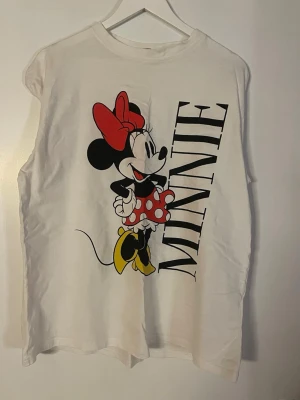 Vit Minnie Mouse t-shirt i bomull - Vit t-shirt med stort tryck av Minnie Mouse och texten 'MINNIE' på framsidan. T-shirten är ärmlös och har en avslappnad passform. Perfekt för dig som gillar Disney och vill ha en lekfull stil. Tillverkad i mjuk bomull.