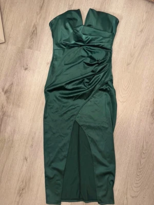 Grön axelbandslös klänning med slits - Mörkgrön axelbandslös klänning med omlott och slits framtill. Klänningen har en figurnära passform och är tillverkad i ett glansigt material som ger en lyxig känsla. Perfekt för dig som vill sticka ut med en elegant look.