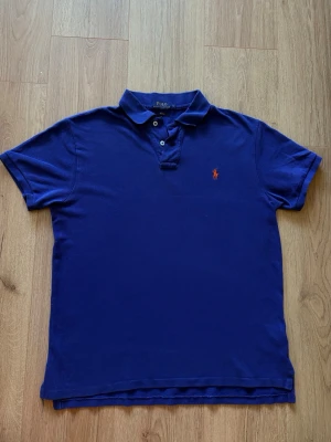 Marinblå pikétröja från Polo Ralph Lauren - Klassisk marinblå pikétröja från Polo Ralph Lauren med korta ärmar och den ikoniska röda logon broderad på bröstet. Perfekt för en clean och stilren look till sommaren