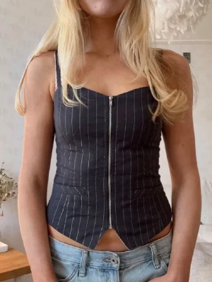 Svart korsett-topp med dragkedja - Snygg svart korsett-topp med tunna axelband och vita vertikala ränder. Toppen har en markerad midja, dragkedja framtill och en liten slits nedtill. Perfekt att styla med jeans för en trendig look.