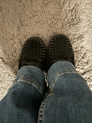 Christian Louboitin skor  - Unika svarta sneakers från Christian Louboutin med coola svarta nitar över hela ovandelen och ikonisk röd sula undertill. Skorna har snörning och är tillverkade i läder för en lyxig känsla. Perfekta för dig som vill sticka ut med din stil.