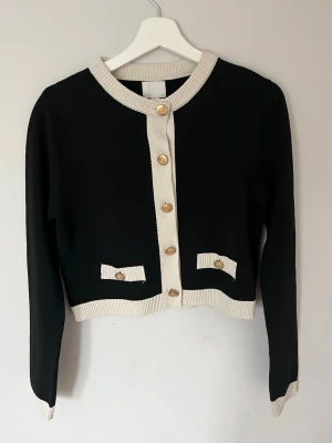 Svart och vit kofta med guldknappar - Stilren svart kofta med vita ribbade kanter och dekorativa guldknappar framtill. Kortare modell med två fejkfickor och rund halsringning. Perfekt för en chic och klassisk look.