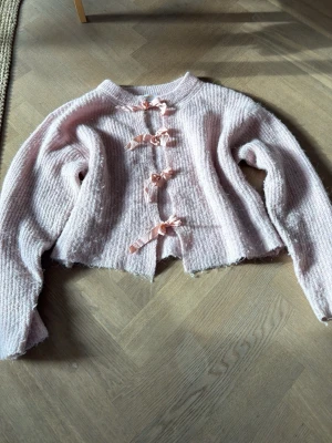 Ljusrosa kofta med rosetter Gina Tricot - Supersöt ljusrosa stickad kofta från Gina Tricot med fyra dekorativa rosetter framtill. Koftan har en croppad passform, lång ärm och är gjord i ett mjukt, fluffigt material. Perfekt för dig som gillar gulliga detaljer och en cozy look.