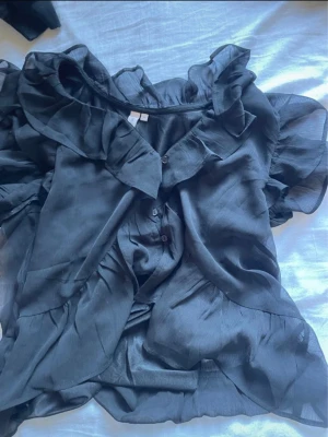 Svart volangblus med knappar - Snygg svart blus i glansigt material med volangdetaljer längs ringningen och ärmarna. Blusen har knäppning framtill och en luftig, flowy passform som ger en romantisk vibe. Perfekt att styla med jeans eller kjol.