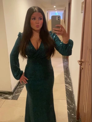 Mörkgrön glittrig långklänning - Snygg mörkgrön långklänning med glittrigt material och v-ringad front. Klänningen har långa ärmar med puffdetaljer och markerad midja som framhäver siluetten. Perfekt för dig som vill sticka ut och känna dig extra glam.