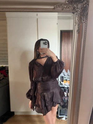 Mörkbrun omlottklänning med volang - Säljer en mörkbrun kort omlottklänning med volangdetaljer och v-ringning. Klänningen har långa, puffiga ärmar och markerad midja med knytband. Materialet har en lätt glans och känns mjukt mot huden. Perfekt för dig som gillar en chic och trendig look. Helt ny med prislapp kvar och hittar inte den på ginas sida längre 