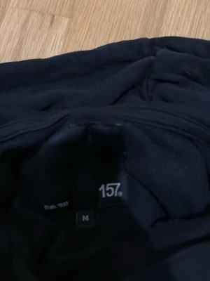 Svart hoodie från 157, storlek M - Klassisk svart hoodie från 157 med huva och dragsko. Hoodien har en stor magficka framtill och ribbade muddar vid ärmslut och nederkant. Perfekt för en chill och avslappnad stil. Tillverkad i mjukt material som känns skönt mot huden.