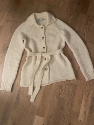 Zara stickad ull kofta - Mysig crèmevit stickad kofta med knappar och matchande bälte i midjan. Koftan har lång ärm, bred krage och ribbade muddar. Perfekt för lager på lager och ger en avslappnad men ändå stilren look. Köpt av en annan tidigare, använd en gång.