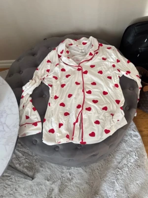 Vit pyjamas med röda hjärtan - Supersöt tvådelad pyjamas i vitt med röda hjärtan över hela setet. Skjortan har krage, knappar framtill och röda passpoaler. Byxorna har resår i midjan och matchande röda detaljer vid benslut. Perfekt för mysiga hemmakvällar.