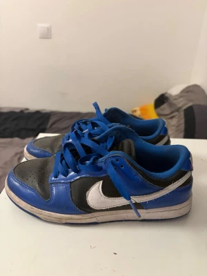 Nike Dunk Low blå och svart - Säljer ett par Nike Dunk Low sneakers i blått och svart med vit swoosh. Skorna har klassisk låg siluett, blå snören och ovandel i läder. Perfekta för dig som gillar streetwear och vill sticka ut med färg. Snygg kontrast mellan blått, svart och vitt.