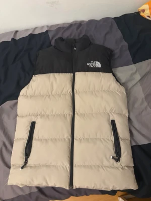Beige och svart dunväst The North Face - Säljer en beige och svart dunväst från The North Face med hög krage och dragkedja framtill. Västen har två svarta dragkedjefickor och logga på bröstet. Perfekt för lager-på-lager och riktigt skön att bära under kyliga dagar.