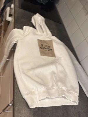 Vit hoodie Burberry med patch - Säljer en vit hoodie från Burberry med en beige patch på bröstet där loggan syns tydligt. Hoodien har klassisk passform med huva och ribbade muddar. Materialet ser ut att vara mjukt och bekvämt, perfekt för en avslappnad stil.