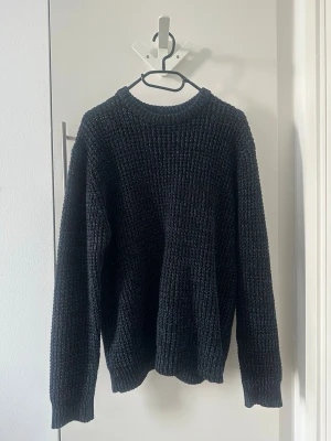 Svart stickad tröja med rund hals - Mysig svart stickad tröja med grov ribbstickning och rund hals. Tröjan har långa ärmar och ribbade muddar vid ärmslut och nederkant. Perfekt för kyliga dagar och enkel att matcha till olika outfits.