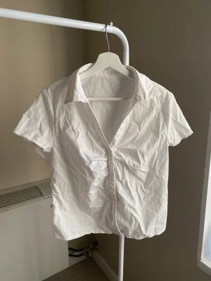 Vit kortärmad skjorta med krage - Klassisk vit skjorta med korta ärmar och krage. Skjortan har knappar framtill och är tillverkad i ett lätt bomullstyg som passar perfekt till sommaren. Enkel och stilren design som funkar till många olika looks.