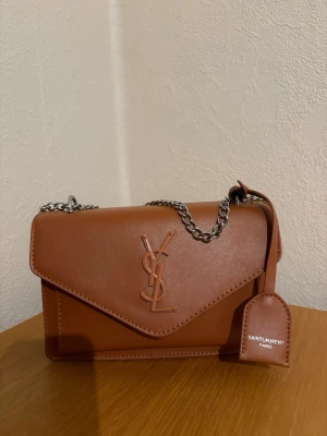 Yves saint laurent väska - Snygg brun väska från Saint Laurent i slätt läder med silverfärgad kedja och YSL-logga på framsidan. Väskan har ett kuvertformat lock och en tag med Saint Laurent Paris. Perfekt för dig som vill ha en trendig och lyxig accessoar.