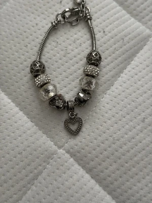 Silverfärgat armband med pärlor och hjärta - Snyggt silverfärgat armband med flera dekorativa pärlor och ett hängande hjärta. Armbandet har detaljer i metall och glasklara pärlor med olika mönster och former. Perfekt accessoar för att lyfta din stil.