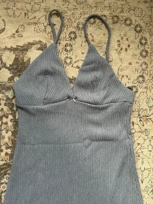 Blå långklänning med smala axelband - Säljer en stilren blå långklänning med smala axelband och v-ringad urringning. Klänningen har en figurnära passform och är tillverkad i ett ribbat material som ger en snygg struktur. Perfekt för dig som gillar minimalistisk och modern stil.