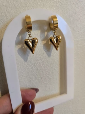 Guldiga hjärtörhängen i rostfritt stål - Snygga örhängen i guldfärgad metall med hängande hjärtan. Hjärtana har en modern, tredimensionell form och örhängena har en chunky ring som fäste. Perfekta för dig som vill ha ett statement-smycke med trendig känsla.
