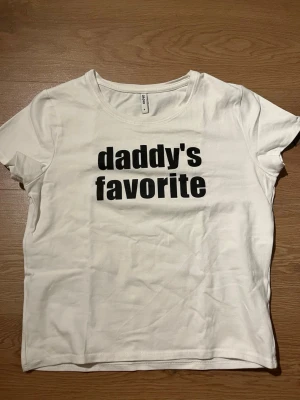 Vit t-shirt från Cropp med tryck - Vit t-shirt från Cropp med svart texttryck 'daddy's favorite' på bröstet. Klassisk rund halsringning och korta ärmar. Tillverkad i mjukt bomullsmaterial för en bekväm känsla. Perfekt för dig som gillar statement-plagg.