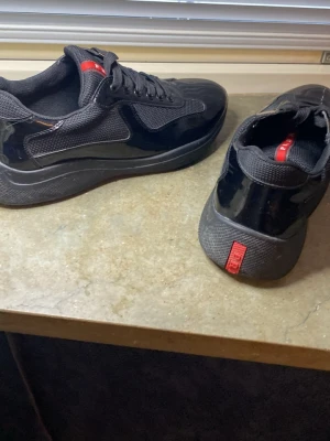 Svarta sneakers - Snygga svarta sneakers med glansig finish och röda detaljer på plösen och hälen. Skorna har en låg profil, svarta snören och en robust sula. Materialet är en mix av mesh och lackat läder för en modern look.