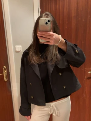 Kort svart trenchcoat - Snygg kort svart trenchcoat med dubbelknäppning och bred krage. Jackan har axelklaffar och dekorativa knappar på axlarna. Perfekt till vår och höst😊