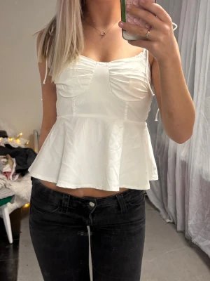 Vit peplum linnetopp - Säljer en söt vit linnetopp med peplumvolang och tunna axelband med knyt. Toppen har markerade kupor och en figurnära passform upptill. Perfekt för sommarens alla tillfällen och lätt att matcha med jeans eller kjol. Små fläckar på baksidan, sista bilden.