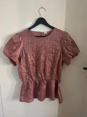 Rosa blus med puffärmar - Söt rosa blus med puffiga korta ärmar och rund halsringning. Blusen har en markerad midja och lätt volang nertill för en feminin look. Materialet har en lätt glansig finish som ger en extra touch. Den är i storlek 170 i barn och från Lindex, men passar dam med storlek XS och S. Den är skrynklig på bilderna men vid en strykning blir den perfekt. Den har en bra stretch och fin glans. Den är nästan aldrig använd! 
