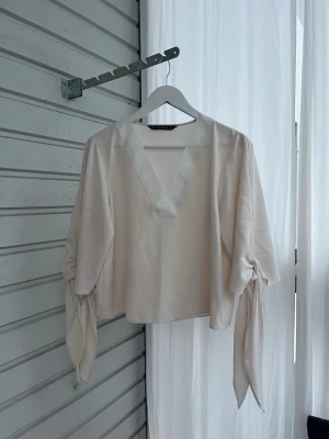 Krämvit blus med knytärmar - Snygg krämvit blus med djup V-ringning och vida långärmar som avslutas med dekorativa knytband. Blusen har en lös passform och är tillverkad i ett lätt, flowy tyg som ger en elegant känsla. Perfekt för att lyfta vilken outfit som helst.