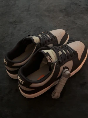 Nike Dunk Low svart och grå - Säljer ett par Nike Dunk Low sneakers i svart och grått med vita detaljer. Klassisk låg siluett med perforerad tåbox och extra skosnören medföljer. Tydlig Nike-logga på hälen och tungan. Perfekta för dig som gillar stilrena och tidlösa sneakers.