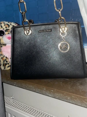 Svart Michael Kors handväska i skinn - Snygg svart handväska från Michael Kors i stilrent skinn med guldfärgade kedjedetaljer och MK-hänge. Väskan har både axelrem och handtag, perfekt för dig som gillar en klassisk och trendig look. Rektangulär form och diskret logga framtill.