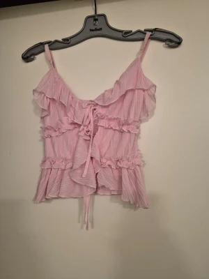 Rosa volangtop med knyt - Supersöt rosa linnetopp med volanger och knyt i ryggen. Toppen har tunna axelband och är tillverkad i ett lätt, luftigt material som passar perfekt till varma dagar. Volangdetaljerna ger en romantisk vibe och toppen är croppad för en trendig look.