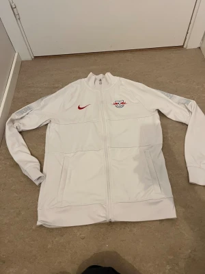 Vit Nike RB Leipzig träningsjacka - Säljer en vit träningsjacka från Nike med RB Leipzig-logga på bröstet och röd Nike-swoosh. Jackan har hel dragkedja, ståkrage och två fickor framtill. Tillverkad i Dri-FIT-material som andas och passar perfekt till träning eller uppvärmning.