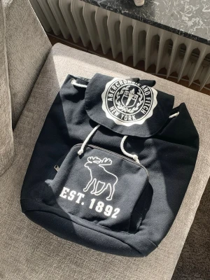 Svart ryggsäck Abercrombie & Fitch - Snygg svart ryggsäck från Abercrombie & Fitch med vitt tryck och logga på locket. Framtill finns en ficka med dragkedja och en stor vit älg samt texten EST. 1892. Ryggsäcken har vita snören för stängning och är gjord i slitstark canvas.