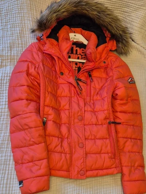 Röd pufferjacka från Superdry - Säljer min svin snygga röda dunjacka från Superdry med avtagbar huva och fuskpäls. Jackan har dragkedja, tryckknappar, flera fickor och coola detaljer på ärmarna. Köpt år 2000 i USA och en ny sådan jacka kostar upp mot 1200. Perfekt för kalla vinterdagar där du förutom kommer se väldigt cool ut kommer vara varm. Klassisk Superdry-logga på ärmen. Skriv för frågor🌸 (är liten i storlek då jag oftast har s-m på jackor men denna jacka har storlek L)