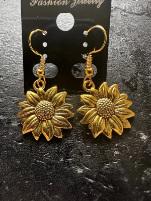 Solrosformade örhängen i guldton - Säljer ett par snygga örhängen i form av solrosor i guldtonad metall. De har krokar för enkel användning och detaljerade blad och mittparti som ger en cool och unik look. Perfekta för dig som vill sticka ut med accessoarer med blommig design.