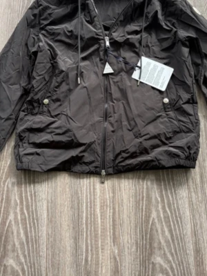 Svart vindjacka f - Snygg svart vindjacka med huva och dragkedja framtill. Jackan har två fickor med tryckknappar och elastiska muddar vid ärmslut och nederkant. På huvan finns en randig detalj i rött och vitt. Tillverkad i lätt, glansigt material.