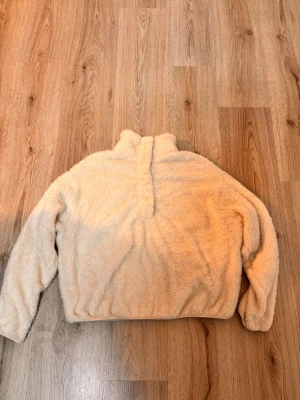 Vit teddytröja med tryckknappar - Mysig teddytröja i vit med hög krage och tryckknappar framtill. Tröjan har en fluffig och mjuk yta som ger en avslappnad look. Perfekt för kyliga dagar när du vill vara både varm och trendig.