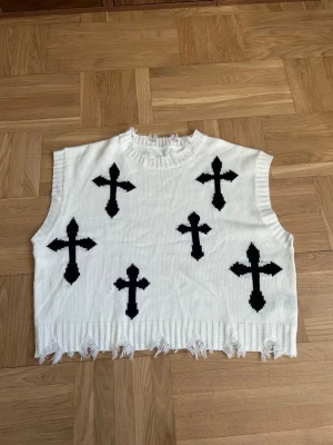 Vit stickad väst med svarta kors - Cool vit stickad väst med svarta korsmönster på framsidan och råa, fransiga kanter. Ärmlös och med ribbad halsringning. Perfekt för att skapa en edgy look. Materialet är mjukt och stretchigt.