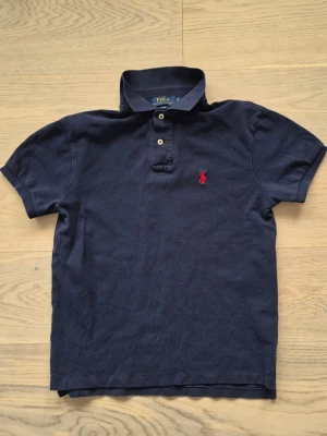 Mörkblå Ralph Lauren Pikétröja - Blå Ralph Lauren Pikétröja med rött emblem. Storlek S. Har något rött märke på baksidan av tröjan men inget man tänker på. (Sista bilden) Annars I fint skick.  Axelbredd: 42cm Längd: 59cm