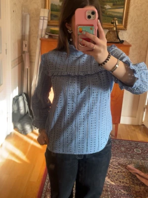 Blå broderad blus med volang - Söt blå blus med broderade hålmönster och volangdetalj över bröstet. Blusen har hög krage med knapp i nacken och långa puffiga ärmar med smock vid ärmslut. Perfekt för dig som gillar romantisk och trendig stil.