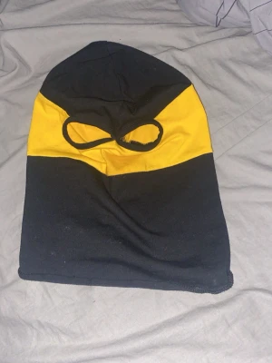 Svart och gul balaclava med hål som är aik  - Säljer en svart och gul balaclava med två ögonhål. Tillverkad i mjukt tyg, perfekt för dig som vill ha en cool och funktionell huvudbonad. Färgerna är tydligt uppdelade med svart nedtill och upptill samt en gul sektion över ögonen. Sim är aik 