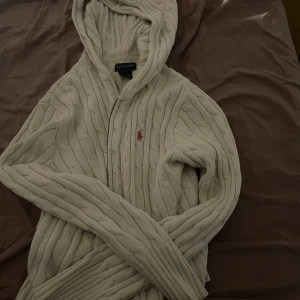 Vit stickad hoodie från Ralph Lauren - Säljer en vit stickad hoodie från Ralph Lauren med dragkedja och det klassiska rosa logobroderiet på bröstet. Tröjan har långa ärmar och en mysig huva, perfekt för kyliga dagar.