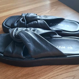 Svarta sandaler från DI NUVO - Säljer ett par svarta sandaler från DI NUVO med breda korslagda remmar i mjukt skinnliknande material. Sandalerna har en chunky sula och öppen tå, perfekt för en avslappnad och trendig look.