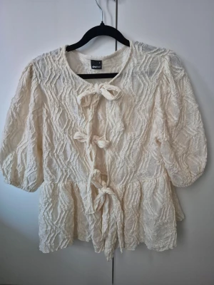 Krämvit blus med knyt från Gina Tricot - Supersöt krämvit blus från Gina Tricot med strukturmönster och tre dekorativa knyt framtill. Blusen har rund halsringning, puffiga trekvartsärmar och ett lätt vågigt fall. 